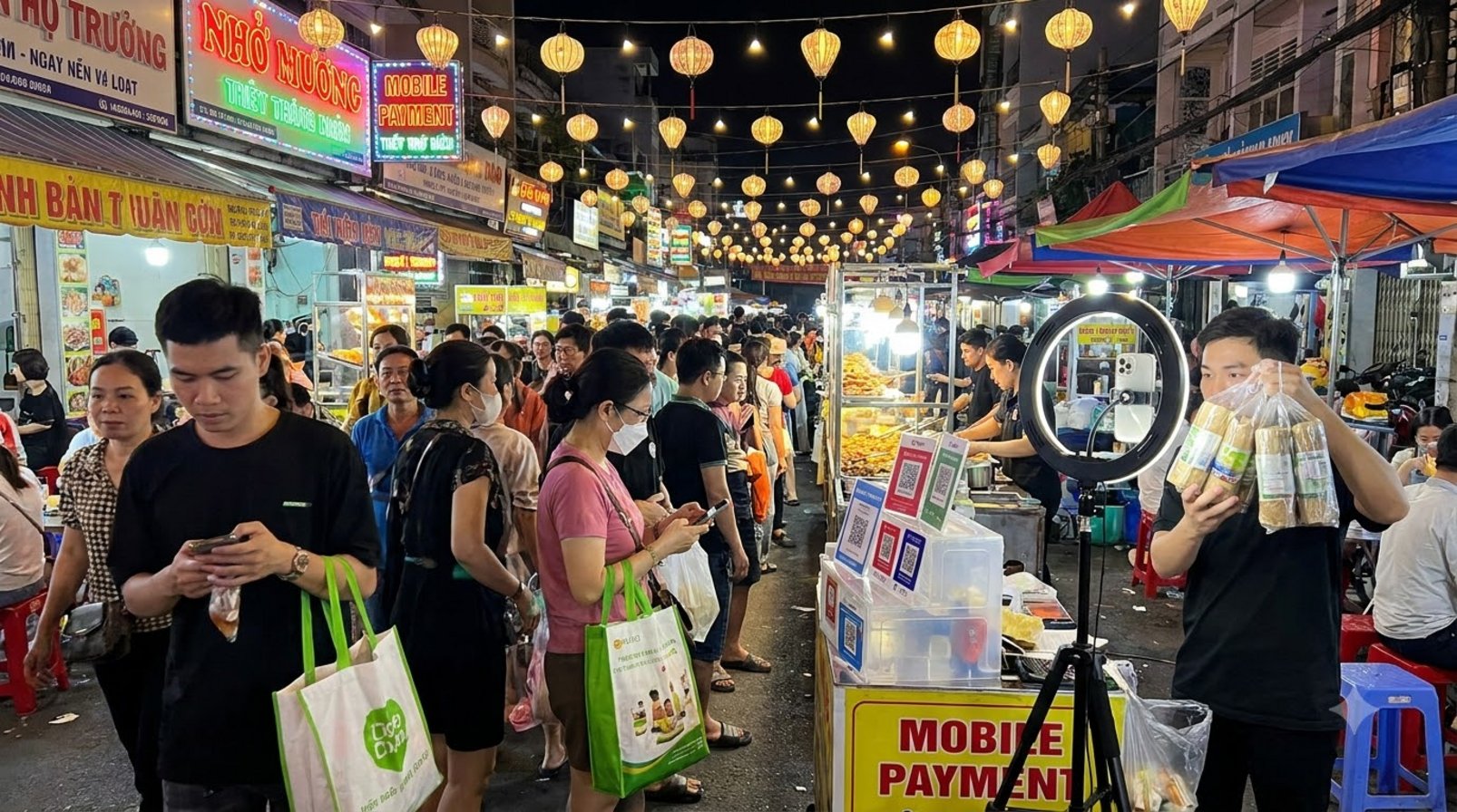 Người mua dạo chợ đêm Việt Nam qua quầy thanh toán, đèn livestream và gian hàng đông khách
